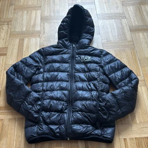 Svart dunjacka EA7 Emporio Armani - Svart dunjacka från EA7 Emporio Armani med huva och quiltad design. Jackan har dragkedja framtill och logga på bröstet. Perfekt för kyliga dagar och riktigt snygg till streetwear-stilen.