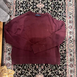 Vinröd sweatshirt från Polo Ralph Lauren - Säljer en vinröd sweatshirt från Polo Ralph Lauren i storlek L. Tröjan har rund halsringning, långa ärmar och klassisk broderad logga på bröstet. Tillverkad i mjuk bomull, perfekt för en avslappnad stil. Endast en liten vit prick på armen