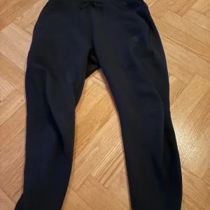 Svarta Nike tech byxor  - Svarta mjukisbyxor från Nike med dragsko i midjan. Perfekta för chill eller träning, klassisk look och bekväm passform. Snygg och enkel design utan synliga detaljer eller mönster.