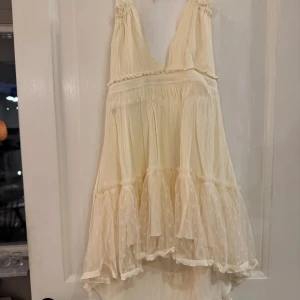 Ljus cremefärgad volangklänning med spets detaljer  - Supersöt ljus cremefärgad klänning med volanger och omlottkänsla. Djup v-ringning och smala axelband, som du kan knyta lite som du vill. Nederdelen har lager av transparent tyg som ger extra volym. Perfekt för sommarens alla tillfällen!