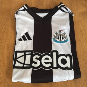 Newcastle United fotbollströja Isak 14 - Snygg Newcastle United fotbollströja med svarta och vita ränder, klubbmärke på bröstet och sponsorlogga 'sela' framtill. På ryggen står det 'Isak' och nummer 14. Klassisk V-ringning och korta ärmar.
