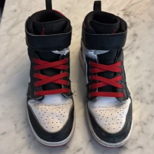Nike Air Jordans med röd snörning - Säljer ett par klassiska Nike Air Jordans med svart och vitt läder, röda detaljer och röd snörning. Skorna har hög modell med kardborreband upptill och ikonisk Air Jordan-logga på plösen. Perfekta för dig som gillar streetwear och sneakers med attityd. Storlek 39