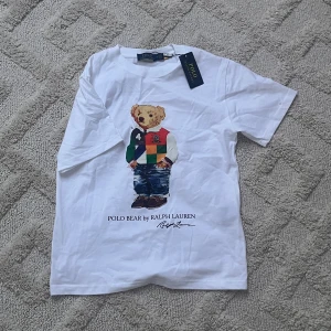 Vit t-shirt med Polo Bear från Ralph Lauren - Helt ny snygg och klassisk T-shirt från Polo Ralph Lauren med det ikoniska “Polo Bear”-motivet framtill. Björnen är klädd i en färgglad rugbytröja och jeans – en lekfull och stilren design som passar perfekt för både vardag och avslappnade tillfällen. T-shirten är i 100 % bomull för en mjuk och bekväm känsla. Helt ny med taggar kvar. En tidlös favorit för dig som gillar kvalitet och karaktär i garderoben