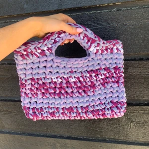Handgjord virka väska i lila och rosa - Superfin virkad väska i lila, rosa och vitt. Väskan har ett chunky, virkat utseende och ett handtag upptill. Perfekt för att addera färg till din outfit och rymmer det viktigaste du behöver ha med dig. Väskan är 30cm bred och 23 vem hög