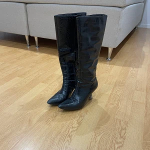 Svarta höga stövlar med krokodilpräglat mönster - Säljer ett par svarta höga stövlar med krokodilpräglat mönster och spetsig tå. Skorna har en medelhög klack och är perfekta för att lyfta vilken outfit som helst.