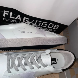 Vita sneakers från Golden Goose Superstar - Golden Goose Superstar sneakers i vitt skinn med grå snörning och silverfärgad stjärndetalj på sidan. Skorna har svart detalj vid hälen och platt sula. Klassisk och stilren modell med rund tå.