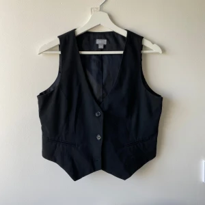 Svart kostymväst med knappar - Klassisk svart väst med två knappar framtill och v-ringning. Västen har fickdetaljer och knytband i ryggen för justerbar passform. Storlek L men passar M också. 