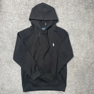 Svart hoodie från Polo Ralph Lauren - Säljer en svart hoodie från Polo Ralph Lauren med klassisk vit broderad logga på bröstet. Tröjan har huva med dragsko och en stor magficka framtill. Perfekt för en avslappnad och stilren look. Pris kan diskuteras!