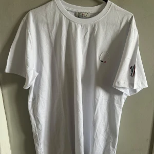 Vit t-shirt från Moncler - Säljer en klassisk vit t-shirt från Moncler med broderad logga på bröstet och diskret Moncler-detalj på ärmen. T-shirten har rund halsringning och är tillverkad i mjuk bomull. Perfekt för en clean och stilren look.