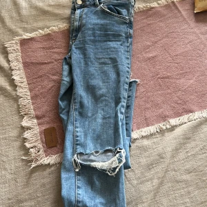 Blå ripped jeans från Never Denim - Snygga ljusblå jeans från Never Denim med slitning och hål vid knät för en trendig look. Klassisk femficksmodell med knapp och dragkedja. Perfekta för dig som gillar en avslappnad och cool stil.