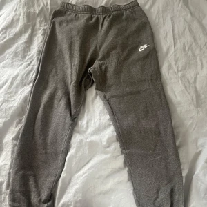 Grå mjukisbyxor från Nike - Gråa mjukisbyxor från Nike med vit logga på vänster lår. Byxorna har resår i midjan och är tillverkade i mjukt bomullsmaterial. Perfekta för chill eller träning och har en klassisk, avslappnad passform.