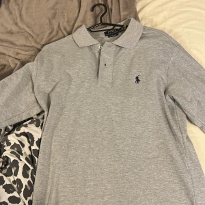 Grå långärmad pikétröja Polo Ralph Lauren - Snygg grå långärmad pikétröja från Polo Ralph Lauren i custom slim fit. Klassisk krage, knäppning framtill och mörkblå broderad logga på bröstet. Perfekt för dig som gillar stilren och enkel design. Tillverkad i mjuk bomull. Pris går att diskuteras vid snabbt köp