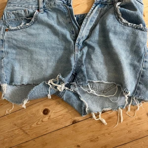 Ljusblå jeansshorts med fransar - Säljer ett par ljusblå jeansshorts med råa, slitna kanter och fransar. Klassisk femficksmodell med dragkedja och knapp. Perfekta för en avslappnad och trendig sommarstil.