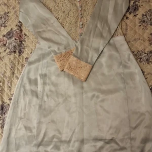 Hemmasydd klänning med spetsdetaljer - Säljer en elegant ljusblå klänning med långärmade ärmar och vackra spetsdetaljer på överdelen och vid ärmsluten. Klänningen har knappar och en figurnära passform. Perfekt för speciella tillfällen. Storleken är ungefär S. Den är en hemmasydd klänning. 