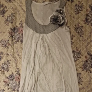 Vit ärmlös tröja med grå krage från Promod - Säljer en vit ärmlös tröja från Promod med en grå prickig krage och dekorativa tygblommor på axeln. Klänningen har en enkel, rak passform och är tillverkad i ett lätt material, perfekt för varma dagar.