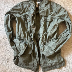 Olivgrön jacka med knappar och dragkedja - Säljer en olivgrön jacka med både knappar och dragkedja framtill. Jackan har flera fickor, markerad midja och klassisk krage. Storlek 40 men är justerbar och passar även mig som har S.