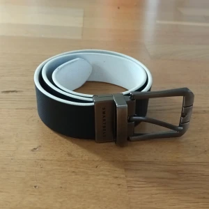 Svart och vitt vändbart bälte från SmartBelt - Stilrent vändbart bälte från SmartBelt med ena sidan svart och andra sidan vit. Bältet har en modern metallspänne med märkets logga ingraverad. Perfekt för att matcha olika outfits och enkelt att justera.