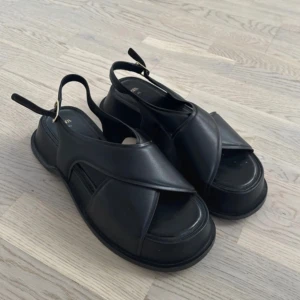 Svarta sandaler från Zara - Säljer ett par svarta sandaler från Zara med breda korslagda remmar och öppen tå. Skorna har en platt sula och justerbart spänne vid hälen. Perfekta för varma dagar och enkel att matcha med olika outfits. Storlek 40, inga defekter 🩷