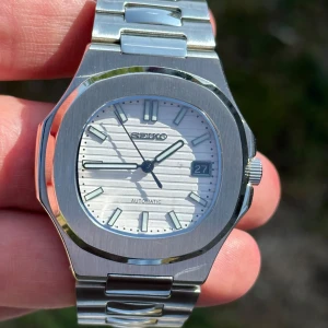 Seiko mod automatisk klocka ”nautilus” - Snygg Seiko automatisk klocka med silverfärgat armband i rostfritt stål och vit urtavla med horisontella ränder. Klockan har datumfönster vid kl 3 och transparent baksida som visar urverket. Modern design med robust länkarmband. Vit urravla, svart datum och safirglas.