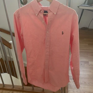 Rosa skjorta från Ralph Lauren Slim Fit - Snygg rosa skjorta från Ralph Lauren i slim fit-modell. Klassisk button-down krage, långa ärmar och broderad logga på bröstet. Skjortan är tillverkad i bomull och har knappar framtill. Perfekt för dig som gillar stilrena och fräscha plagg.