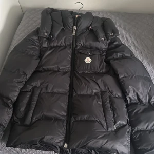 Svart dunjacka från Moncler - Säljer en svart dunjacka från Moncler med hög krage och dragkedja framtill. Jackan har en bröstficka med Moncler-logga och två sidofickor. Klassisk pufferstil med quiltad design och lång ärm.