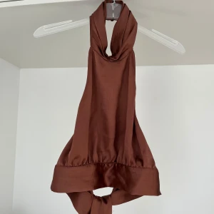 Brun halterneck topp - Säljer en snygg brun halterneck topp med knyt i ryggen. Toppen har en croppad passform och är perfekt för varma dagar. Tillverkad i ett mjukt sidenmaterial som känns skönt mot huden.