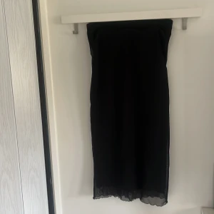 Svart axelbandslös långklänning - Säljer en svart axelbandslös långklänning med enkel och stilren design. Klänningen har en rak passform och är tillverkad i ett lätt, syntetiskt material som faller snyggt. Perfekt för dig som gillar minimalistisk stil.