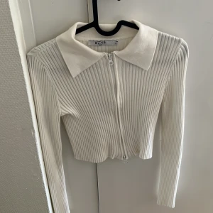 Vit ribbad kofta med dragkedja från NA-KD - Säljer en vit ribbad kofta från NA-KD med dragkedja framtill och stor krage. Tröjan är croppad och har långa ärmar. Perfekt att styla till olika outfits!