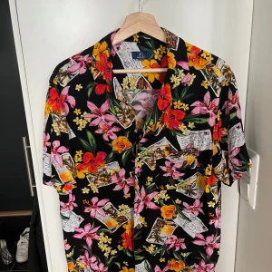 Klassisk hawaii-skjorta  - Färgglad skjorta med svart bas och stora blommor. Skjortan har korta ärmar, klassisk krage och knäppning framtill. Tillverkad i 100% viskos för en luftig känsla. Perfekt för dig som vill få extra semesterkänsla i sommar!  