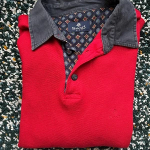 Röd pikétröja från Sand med blå krage - Röd pikétröja från Sand med kontrasterande blå krage och knäppning framtill. Tröjan har lång ärm, broderad logga på bröstet och mönstrat tyg på insidan av kragen. Tillverkad i bomull för en skön och avslappnad känsla.