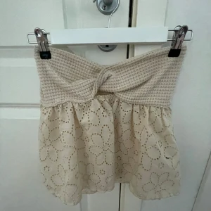 Beige tubtopp - Supersöt beige tubtopp med virkad, twistad midja och broderad nederdel i blommigt mönster. Kjolen har en luftig känsla och passar perfekt till varmare dagar. Den stickade midjan ger en unik och trendig look. 