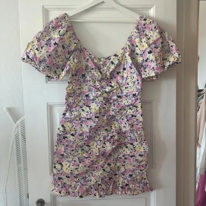 Blommig kortklänning med puffärmar - Superfin kortklänning med blommigt mönster i rosa, lila, gul och svart. Klänningen har puffiga korta ärmar, v-ringning och volangdetalj nertill. Perfekt för sommarens alla tillfällen!