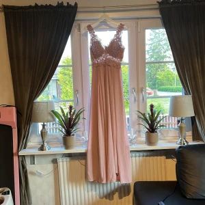 Långklänning med spets och blommiga detaljer - Säljer en elegant ljusrosa långklänning med vackert blommigt spetsliv och breda axelband. Klänningen har en flowy kjol och transparenta partier upptill. Perfekt för bal eller fest! 🌸