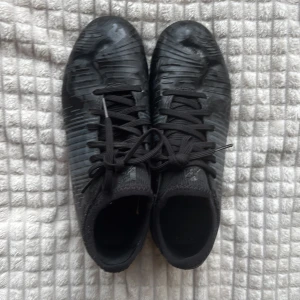 Svarta fotbollsskor med snörning - Säljer ett par svarta fotbollsskor från Puma med snörning och mönstrad ovandel. Skorna har rund tå och fasta dobbar för spel på gräs. Perfekta för dig som vill ha stilrena och funktionella skor på planen.💕