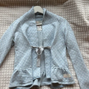 Ljusblå mönstrad cardigan jacka från Odd Molly - Superfin ljusblå cardigan jacka från Odd Molly med vitt diskret mönster. Jackan har lång ärm, knytband i midjan och två fickor fram. Broderad detalj på ryggen och mjukt material som känns skönt mot huden.