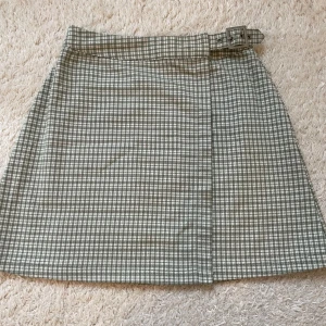 Rutig kortkjol - Grön och vit rutig kortkjol omlott med spänne i midjan. Kjolen har en klassisk A-linjeform. Tidlös design som funkar till många olika looks. Från brandy melvill därav onesize men passar xs. Lite nopprig i tyget på vissa ställen. 