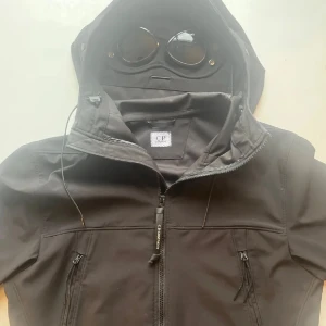 Svart Goggle Jacket från C.P. Company - Säljer en svart Goggle Jacket från C.P. Company. Jackan har en unik design med inbyggda glasögon i huvan och dragkedja framtill. Tillverkad i ett stretchigt soft shell-material som är perfekt för varierande väder. Två fickor med dragkedja på framsidan ger extra funktionalitet.