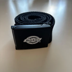 Svart bälte från Dickies - Snyggt svart bälte från Dickies med en stilren spänne i metall. Perfekt för att ge en cool touch till din outfit. Bältet är justerbart och passar de flesta storlekar.