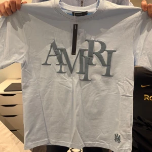 Ljusblå t-shirt från Amiri - Säljer en stilren ljusblå t-shirt från Amiri med ett stort, upphöjt AMIRI-tryck på framsidan. T-shirten är kortärmad och har en rund halsringning. Perfekt för en avslappnad och trendig look. strl L