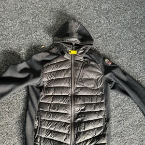 Svart Parajumper med huva - Snygg svart pufferjacka från Parajumpers med huva och dragkedja. Jackan har långa ärmar och en quiltad design. Perfekt för kyligare dagar med sin stilrena look och praktiska detaljer. Nypris 4500kr, Strl Large