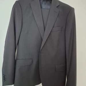 Mörk marinblå kostym med kavaj och byxor - Oscar Jacobsson. Elegant mörk marinblå kostym med kavaj och matchande byxor. Kavajen har klassisk design med knappar och fickor. Perfekt för formella tillfällen. Helt nytt, aldrig använd. Färgkod 210. Style 3050, Kvalitet 4158. Byxa storlek 100, kavaj storlek 104