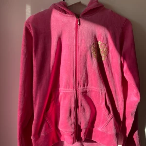 Rosa hoodie från Juicy Couture - Säljer en snygg, gammal version rosa hoodie från Juicy Couture med dragkedja och huva. Den har ett guldglittrigt emblem på bröstet och två praktiska fickor framtill. Perfekt för en avslappnad stil med en touch av glamour. Står att det är storlek XXL men passar mig perfekt då jag har storleken S/M (pris kan diskuteras)