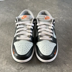Nike Dunk i svart och vit - Snygga Nike Dunk sneakers i svart och vit med orange detaljer. Skorna har en klassisk design med snörning och rund tå. Perfekta för en stilren look. Är i väldigt bra skick och har vart använda ett par gånger