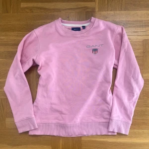 Rosa sweatshirt från GANT - Säljer en snygg rosa sweatshirt från GANT med broderad logga och emblem på bröstet. Tröjan har rund hals och långa ärmar, perfekt för en avslappnad stil. Passar bra till både jeans och kjol. 🌸