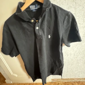 Svart pikétröja från Ralph Lauren - Säljer en klassisk svart pikétröja från Ralph Lauren med den ikoniska loggan broderad på bröstet. Tröjan har en klassisk passform med knappar vid kragen. Perfekt för en stilren look.