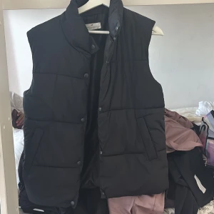 Svart dunväst från H&M - Säljer en stilren svart dunväst från H&M. Västen har en hög krage och knäpps med knappar framtill. Perfekt för lager-på-lager under kyliga dagar. Passar bra till både casual och mer uppklädda tillfällen. Använd flera gånger men i bra skick barn strl 12-14 år