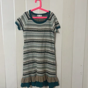 Randig stickad klänning med volang - Charmig stickad klänning med randigt mönster i svart, beige och grön. Den har korta ärmar och en söt volangdetalj längst ner. Perfekt för en avslappnad stil.