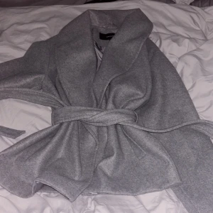Grå kappa från Vero Moda - Snygg grå kappa från Vero Moda med ett elegant bälte i midjan. Den har en stilren design med bred krage och är perfekt för kyligare dagar. Fodrad med ett glansigt material för extra komfort.