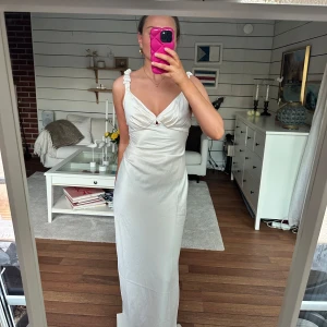 Vit långklänning från Adoore - Elegant vit långklänning med smala axelband och volangdetaljer. Klänningen har en v-ringad design och en åtsittande passform som framhäver figuren. Perfekt för speciella tillfällen. Klänningen är helt ny och enbart provad.