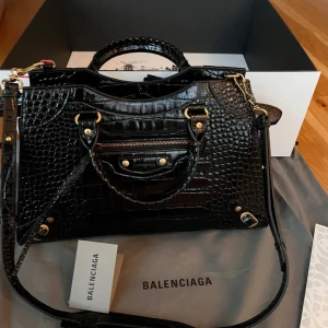 Svart axelväska från Balenciaga - Snygg svart axelväska från Balenciaga i krokodilmönstrat skinn. Väskan har en justerbar axelrem och flera detaljer i guld, inklusive dragkedja och nitar. Perfekt för att ge din outfit en lyxig touch.
